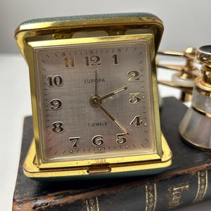 Puede incluir: Un reloj de viaje Europa vintage con una caja dorada y una esfera plateada. El reloj tiene números y manecillas negras, y las palabras "7 JEWELS" y "GERMANY" están impresas en la esfera. El reloj está abierto y descansa sobre un libro oscuro.
