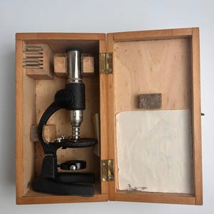 Puede incluir: Un microscopio de metal negro con ocular y lente objetivo plateados, asegurado dentro de una caja de almacenamiento de madera clara. La caja cuenta con soportes internos de madera, una bisagra de latón y un inserto de papel blanco.
