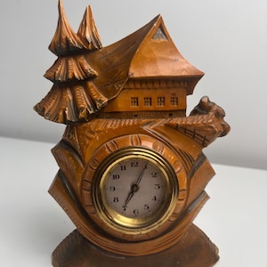 May include: A carved wooden clock shaped like a house with a pine tree on the left side. The clock face is round with a gold-colored metal rim, white background, black numbers from 1 to 12, and two black hands indicating the current time.