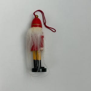 May include: A small wooden Christmas ornament in the shape of a toy soldier. The ornament has a red hat and jacket, yellow pants, and black boots. It has a white beard and a red string for hanging.