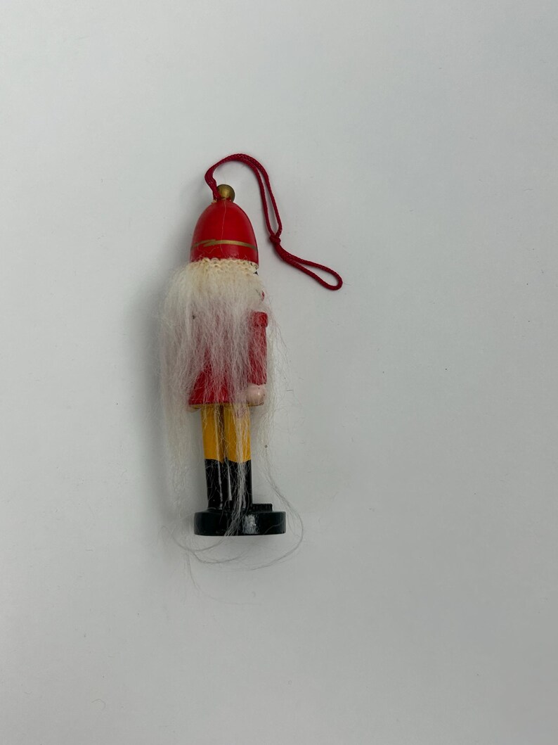 May include: A small wooden Christmas ornament in the shape of a toy soldier. The ornament has a red hat and jacket, yellow pants, and black boots. It has a white beard and a red string for hanging.