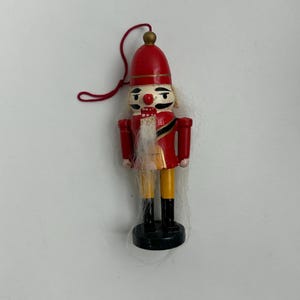 May include: A miniature wooden nutcracker ornament. The ornament features a red hat and jacket, yellow pants, black boots, and a white beard. A red string is attached for hanging. The nutcracker is approximately 4 inches tall.