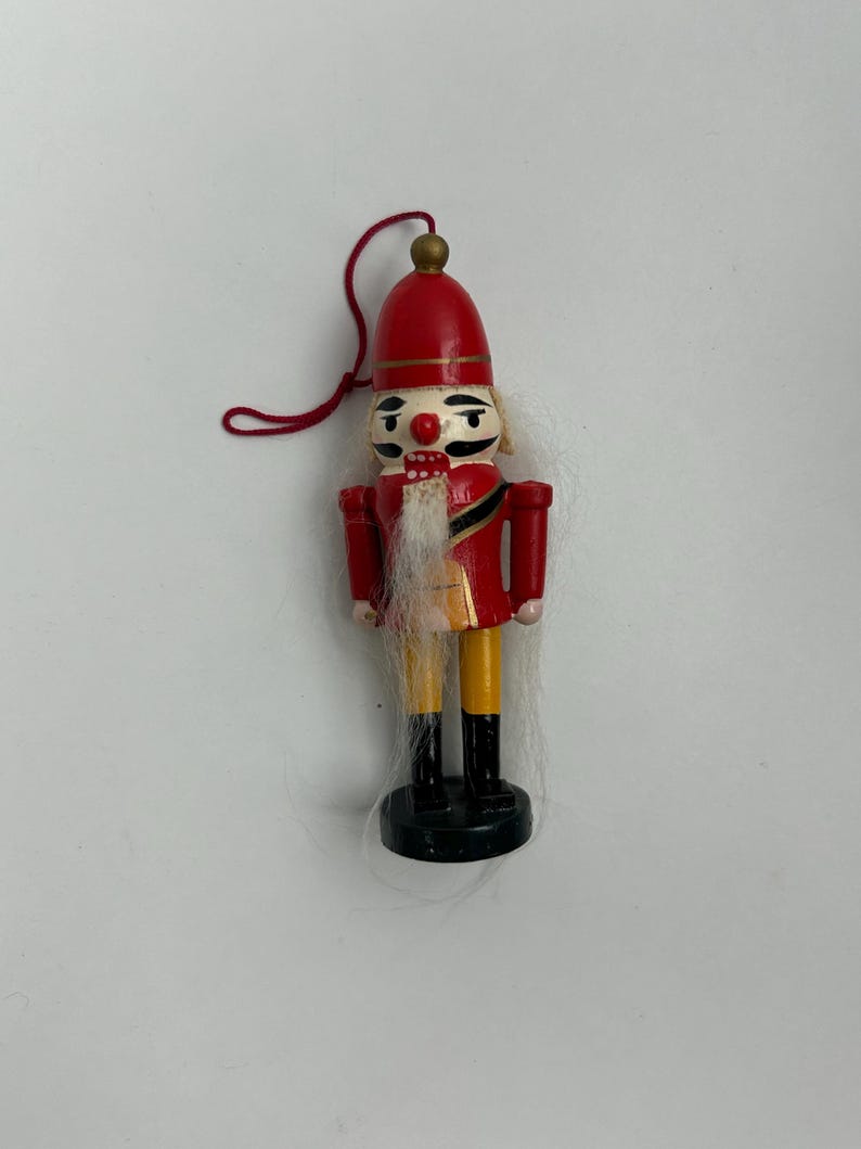 May include: A miniature wooden nutcracker ornament. The ornament features a red hat and jacket, yellow pants, black boots, and a white beard. A red string is attached for hanging. The nutcracker is approximately 4 inches tall.