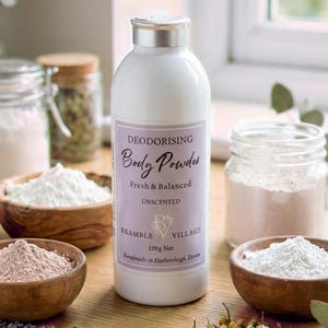 May include: A white bottle of unscented deodorising body powder with a purple label, surrounded by small wooden bowls filled with powder and jars of herbs. The label reads "Bramble Village".