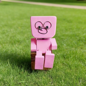 May include: A pink wooden robot figurine with a whimsical design. The robot has a square head with wire detailing for eyes and mouth. It is seated on a small wooden block, with its arms and legs positioned. The background is a blurred green lawn.