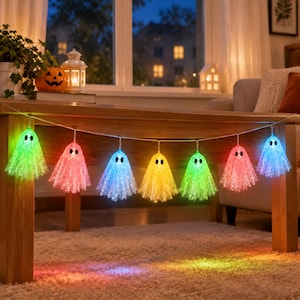 Op de afbeelding: Een lichtsnoer met zeven doorschijnende, spookvormige kapjes in verschillende kleuren, waaronder groen, roze, blauw en geel. Elk spookje heeft twee zwarte, ovale ogen en wordt van binnenuit verlicht, wat een kleurrijke gloed op de vloer werpt.