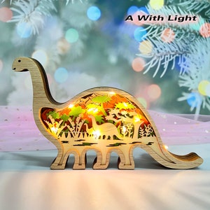 Könnte beinhalten: Eine hölzerne Dinosaurier-Nachtlampe mit einem ausgeschnittenen Design einer Waldszene und Lichterketten im Inneren. Der Dinosaurier steht auf einer weißen Oberfläche. Der Text "A With Light" ist in der oberen rechten Ecke des Bildes sichtbar.