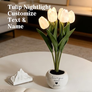 May include: A white ceramic tulip nightlight with six illuminated tulip blooms and green leaves. The nightlight is in a round white pot with the text "I love you mom" and the name "Fiona". The text "Tulip Nightlight Customize Text & Name" is in the image.