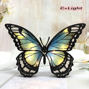 以下が含まれることがあります： 青と黄色のグラデーションデザインと内蔵LEDライトが付いた木製の蝶の壁飾り。蝶は暖色系の白い光で照らされています。画像の右上に「C+Light」というテキストが表示されています。