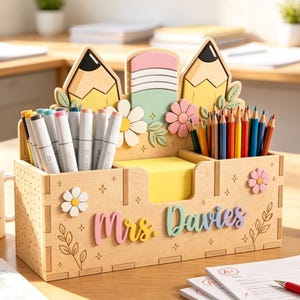 May include: A wooden desk organizer featuring the name Mrs. Davies in colorful 3D lettering. The back panel displays two yellow pencils and a green eraser, accented with flowers. It holds markers, colored pencils, and a stack of yellow sticky notes.