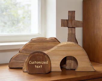 May include: Wooden Easter scene featuring a cross, a tomb with an arched opening, and a mountain-like structure. The tomb has a small, rounded stone with the text "Customized text". The scene is crafted from light brown wood.