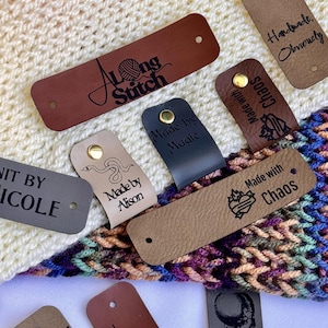 May include: A collection of leather tags in various colors, including brown, black, and gray, with gold rivets. The tags feature phrases like "Along Stitch," "Made by Magic," and "Made with Chaos." Some tags have illustrations, and the background is a knitted fabric.