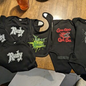 May include: Five black baby onesies with different designs. One onesie says "Rudy" in a white, gothic font. Another onesie says "Goo Goo Ga Ga" with a red dragon graphic. Another onesie says "I What Birdy?" with a black and white bull graphic. A black bib with a green and white splatter design says "Vormit Kebluft".