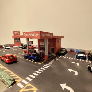 May include: Miniature model of a Speedway gas station with several toy cars parked on the asphalt. The gas station has a red and white canopy with the Speedway logo. The cars are various colors, including red, blue, and white.