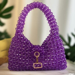 May include: A handcrafted, purple beaded handbag with a structured design. The bag features a rounded handle and a gold-colored clasp. The beads are arranged in a geometric pattern, creating a textured surface. The bag is designed to be carried by hand.