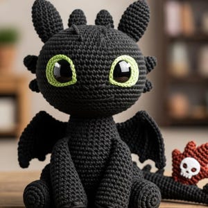 May include: A black crocheted dragon toy with large green-rimmed eyes, small wings, and a friendly expression. The handmade dragon is sitting on a wooden surface. A small red and white skull-shaped toy is next to the dragon.