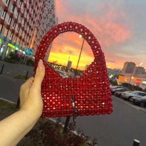 May include: A red beaded handbag with a curved handle and a rectangular body. The bag features a silver-colored clasp and is held against a sunset backdrop. The beads appear to be small and closely packed.