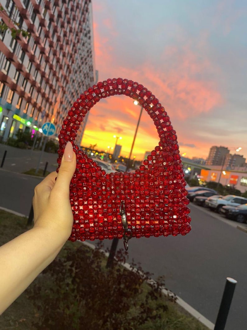 May include: A red beaded handbag with a curved handle and a rectangular body. The bag features a silver-colored clasp and is held against a sunset backdrop. The beads appear to be small and closely packed.