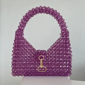 May include: A handcrafted, purple beaded handbag with a rounded handle and a gold-colored clasp. The bag is made of small, translucent beads, giving it a textured appearance. The bag's design is a unique, stylish accessory.