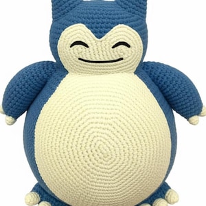 Puede incluir: Un peluche de ganchillo azul y crema hecho a mano. El juguete es un personaje redondo y sonriente con orejas, brazos y pies pequeños. El cuerpo es de color crema y el resto del juguete es azul. Es un Snorlax de la serie Pokemon.