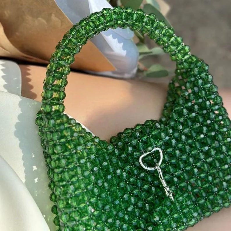 May include: A green beaded handbag with a curved handle and a silver-colored clasp. The bag is made of small, square-shaped beads, giving it a textured appearance. The handle is also beaded, and the bag appears to be a small, stylish accessory.
