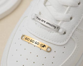 Könnte beinhalten: Weiße Sneaker mit zwei goldenen Schuh-Anhängern. Die Anhänger sind mit dem Text "GO GO GO GO" und "You are amazing!!" graviert.