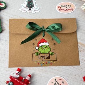 May include: A brown paper gift bag tied with a green satin ribbon. The bag features a Grinch graphic wearing a Santa hat and holding a sign that says "Crystal". A string of colourful lights is below the sign. Christmas themed stickers are scattered around the bag.