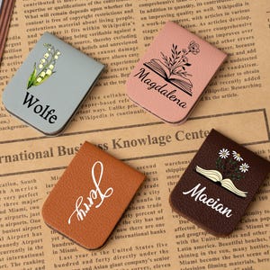 Può includere: Quattro segnalibri in pelle personalizzati. Uno è grigio chiaro con un motivo floreale e il nome "Wolfe". Un altro è rosa con un libro e il nome "Magdalena". Un terzo è marrone con il nome "Jerry". L'ultimo è marrone scuro con margherite e il nome "Maeian".