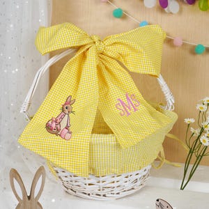 Puede incluir: Una cesta de Pascua de mimbre blanco con forro y lazo de cuadros amarillos. La cesta presenta un diseño de conejito bordado e iniciales con monograma. Un artículo decorativo con temática de Pascua.