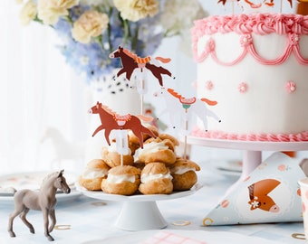 May include: A white cake with pink frosting and horse-themed decorations sits on a pink cake stand. A plate of cream puffs topped with whipped cream and horse-themed toppers sits on a white pedestal. A brown horse figurine sits on a blue and white tablecloth.