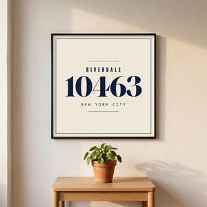 May include: Framed wall art featuring the text "RIVERDALE 10463 NEW YORK CITY" in navy blue on a cream background. The artwork is in a black frame and sits above a small potted plant on a wooden table.