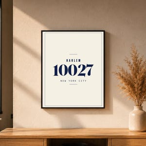 Può includere: Stampa artistica incorniciata con il testo "HARLEM 10027 NEW YORK CITY" in blu navy su sfondo crema. La stampa è in una cornice nera. Un vaso con fiori secchi è a destra. La stampa è su una superficie di legno.