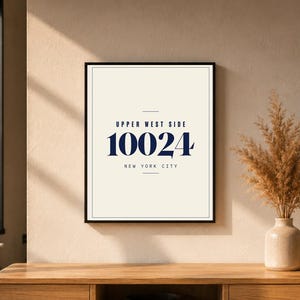 Può includere: Una stampa incorniciata con il testo "UPPER WEST SIDE 10024 NEW YORK CITY" in blu navy su sfondo bianco sporco. La stampa è in una cornice nera ed è esposta su una superficie di legno. Un vaso con piante secche è accanto alla cornice.