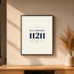 Könnte beinhalten: Ein gerahmter Druck mit dem Text "WILLIAMSBURG 11211 NEW YORK CITY" in Marineblau auf cremefarbenem Hintergrund. Der Druck befindet sich in einem schwarzen Rahmen. Rechts steht eine Vase mit getrockneten Pflanzen.