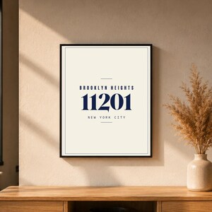May include: Framed print featuring the text "BROOKLYN HEIGHTS 11201 NEW YORK CITY" in navy blue on a cream background. The print is in a black frame and is displayed on a wooden surface. A vase with dried plants is next to the print.
