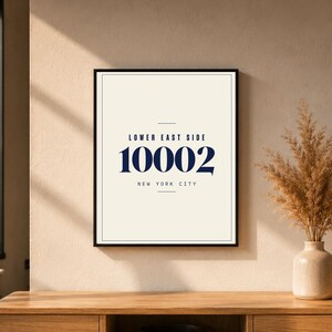 May include: A framed print with the text "LOWER EAST SIDE 10002 NEW YORK CITY" in navy blue on an off-white background. The print is displayed on a wooden surface, with a vase of dried flowers to the right.