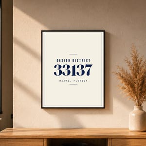 Könnte beinhalten: Gerahmter Kunstdruck mit dem Text "DESIGN DISTRICT" und der Nummer "33137" über "MIAMI, FLORIDA". Der Druck befindet sich in einem schwarzen Rahmen und wird auf einer Holzoberfläche angezeigt. Eine Vase mit getrockneten Blumen ist ebenfalls zu sehen.