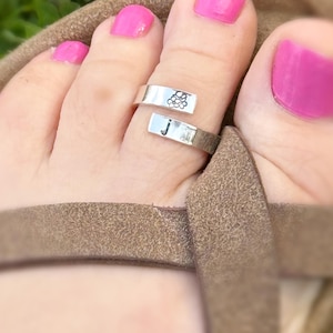 May include: A silver toe ring with a floral design and the letter "J" is worn on a foot with pink nail polish. The foot is in a brown sandal, with green grass in the background.
