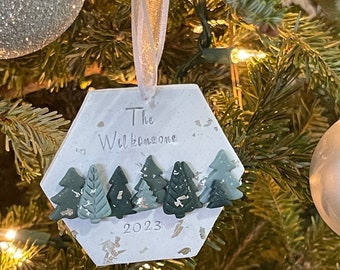 May include: White hexagonal Christmas ornament with the text "The Wilkinsons" and the year "2023". The ornament is decorated with green and gold glitter and features a row of small green Christmas trees.