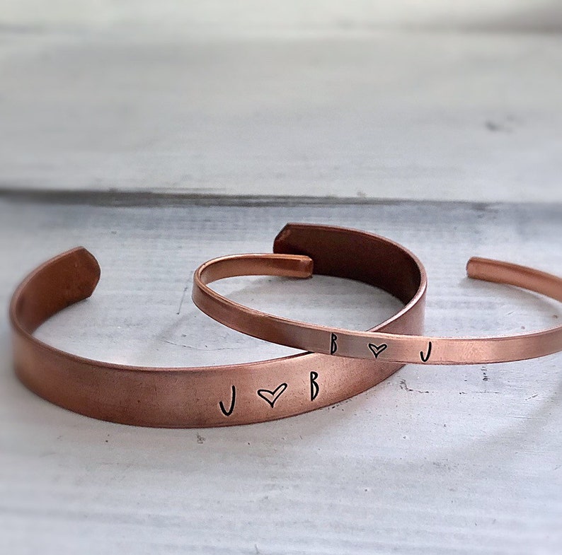 May include: Two copper bracelets with engraved initials and a heart symbol. The bracelets are stacked on top of each other, with the top bracelet reading "B &hearts; J" and the bottom bracelet reading "J &hearts; B".
