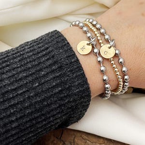 May include: A set of three beaded bracelets. Two bracelets feature silver beads, and one has gold beads. Each bracelet has a gold charm with a letter. One charm has the letter "K", and the other has the letter "C".