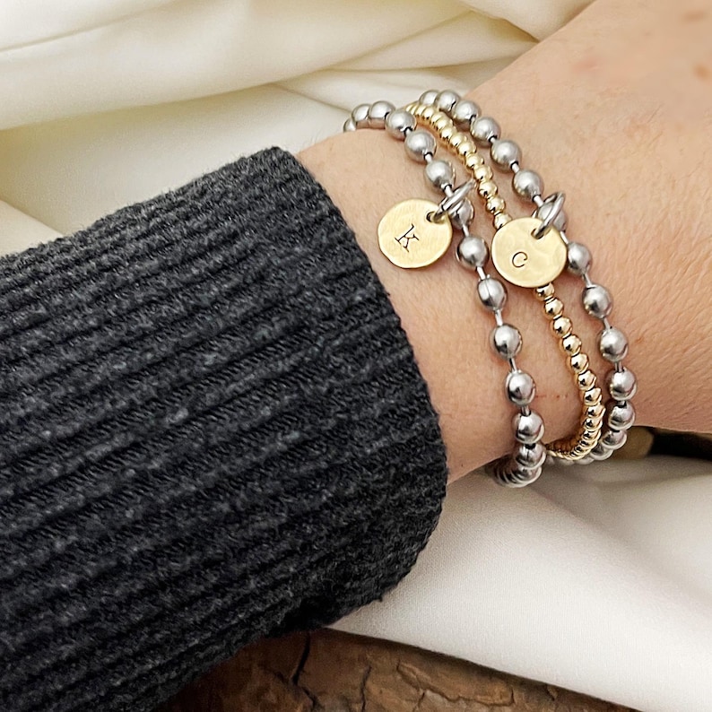 May include: A set of three beaded bracelets. Two bracelets feature silver beads, and one has gold beads. Each bracelet has a gold charm with a letter. One charm has the letter "K", and the other has the letter "C".