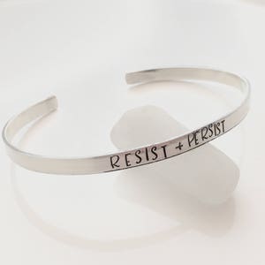 May include: A silver bangle bracelet with the words "Resist + Persist" engraved on it.