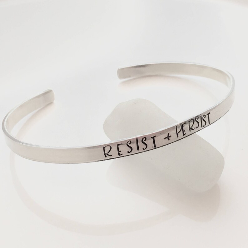 May include: A silver bangle bracelet with the words "Resist + Persist" engraved on it.