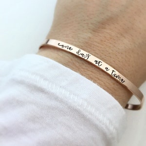 May include: A rose gold bangle bracelet with the inscription "one day at a time".