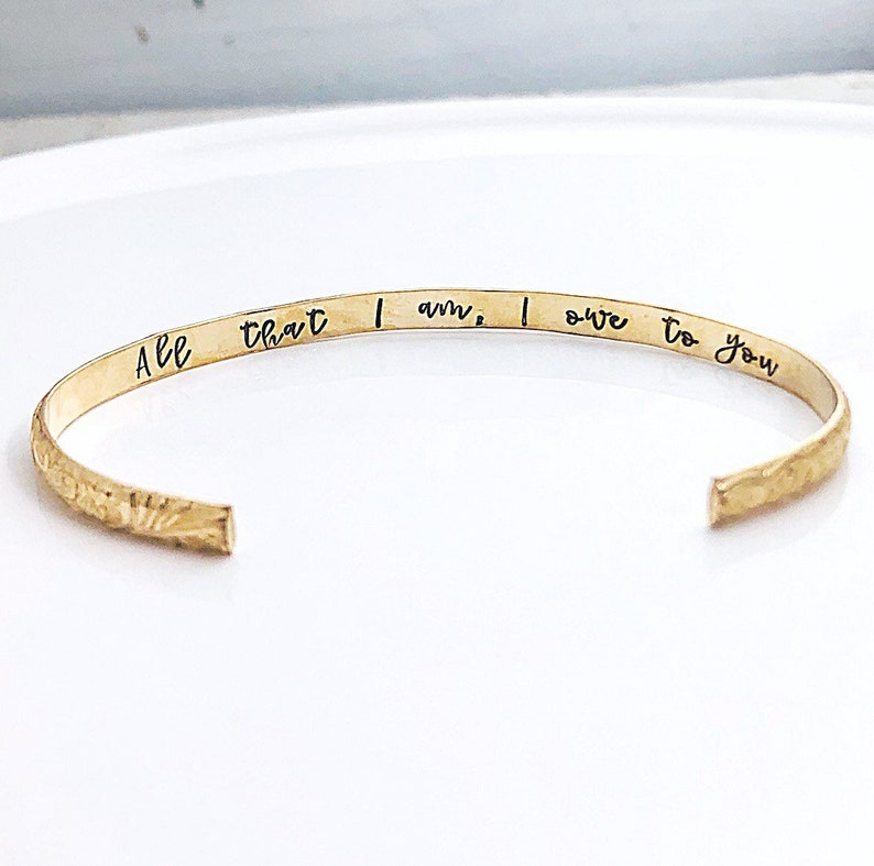 May include: Gold cuff bracelet with the inscription "All that I am, I owe to you".