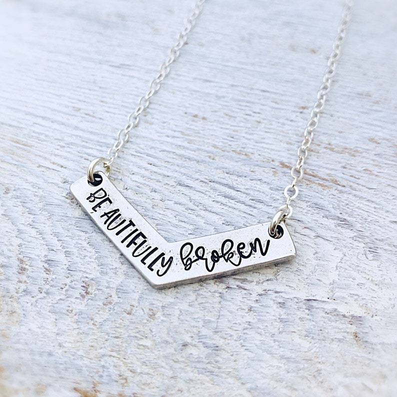K&ouml;nnte beinhalten: Silberne Halskette mit einem Chevron-f&ouml;rmigen Anh&auml;nger, auf dem "Beautifully broken" steht.