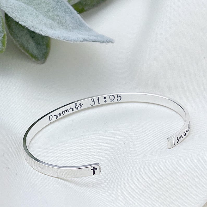 May include: A silver cuff bracelet with the inscription "Proverbs 31:25" and a small cross. The bracelet also has the name "Isabel" engraved on it. The bracelet is displayed on a white surface.