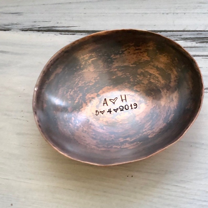 May include: An oval-shaped copper bowl with a hammered, dark patina finish. The interior is stamped with the letters A and H separated by a heart symbol, followed by the date 5, a heart, 4, a heart, and 2019, all in a simple sans-serif font.