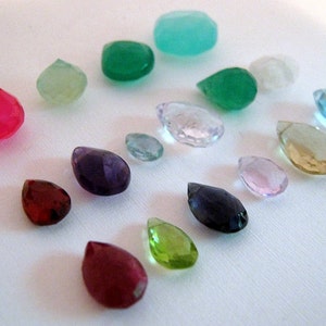 Puede incluir: Una colecci&oacute;n de gemas facetadas en varias formas y colores, incluyendo rojo, verde, morado y azul. Las gemas est&aacute;n dispersas sobre una superficie blanca, mostrando sus diferentes cortes y claridad.